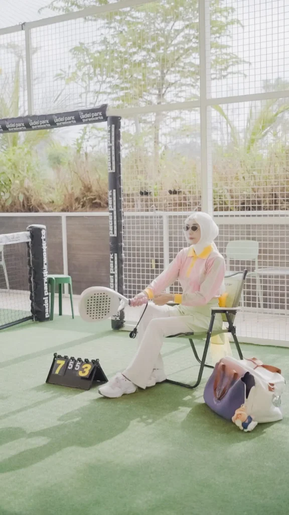 Wanita berhijab, pakaian sporty, berpose duduk, latar padel park.
