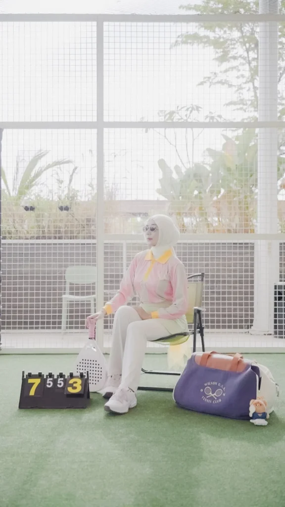 wanita berpose di latar belakang jaring, pakaian santai padel, topi, dan tas tenis.