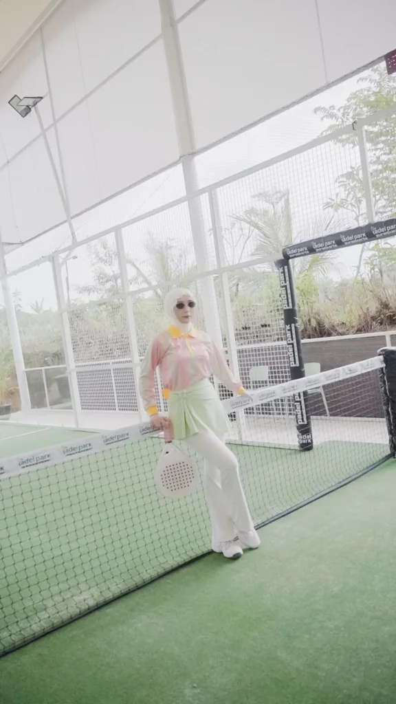 wanita berpose di lapangan padel, hijab, kemeja pink, celana hijau, topi, latar belakang jaring dan