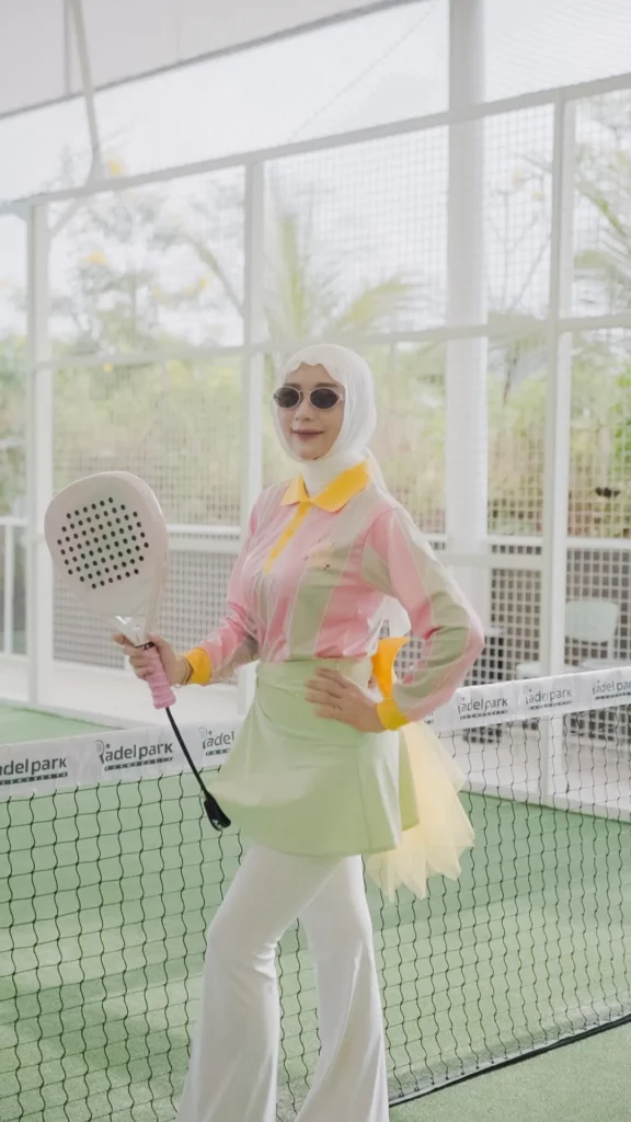Wanita berpose di lapangan padel, hijab, pakaian sporty, latar belakang jaring.