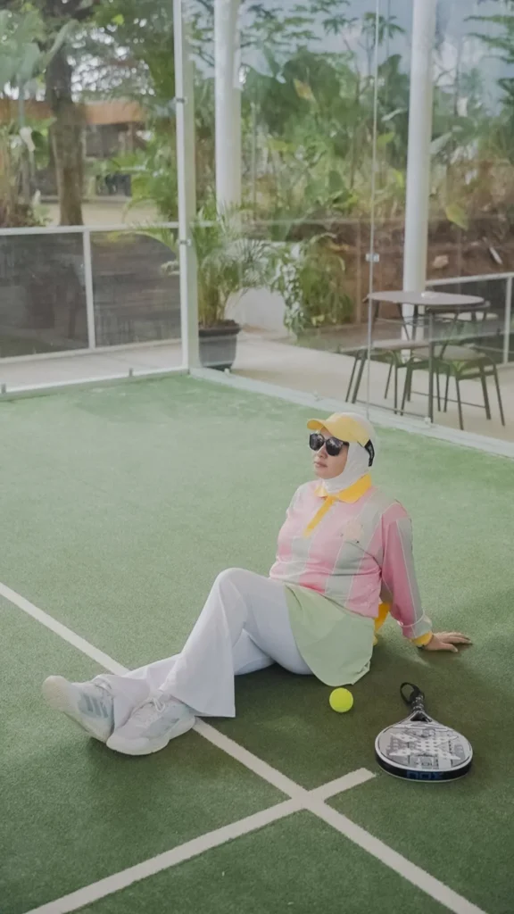 wanita duduk di lapangan padel berpose santai, pakaian modern, latar belakang taman hijau.