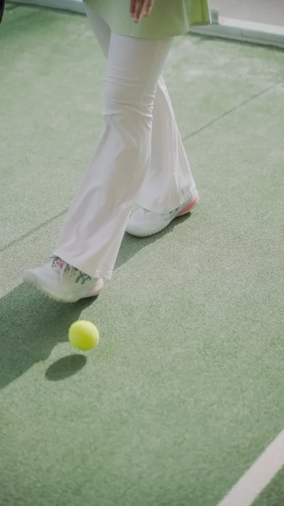 wanita bermain padel dengan celana panjang putih, berpose di lapangan tenis hijau.