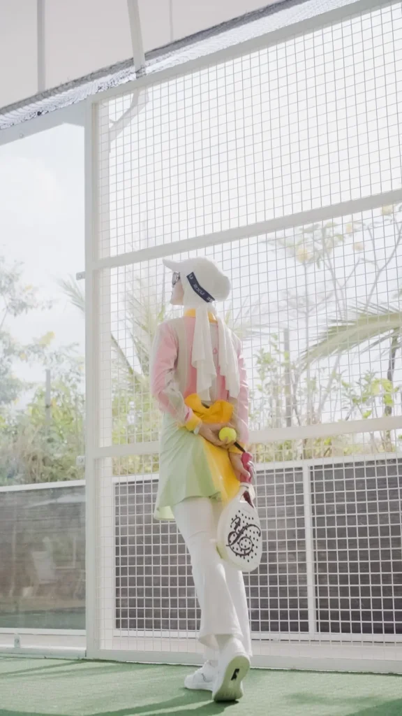 wanita berpose dengan raket padel, pakaian formal, latar belakang pagar.