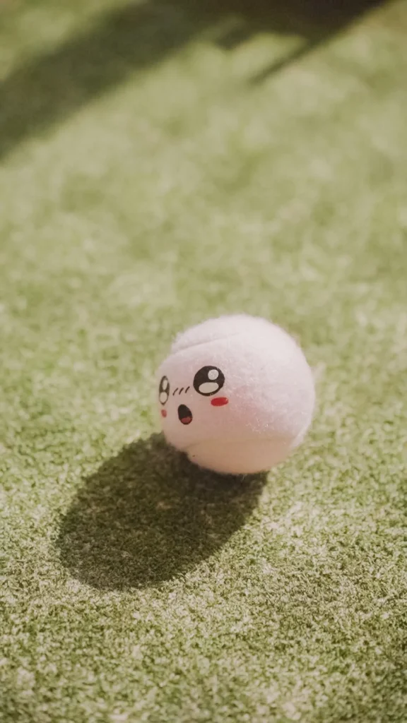 Boneka bola lucu berwajah gembira, duduk di karpet hijau.