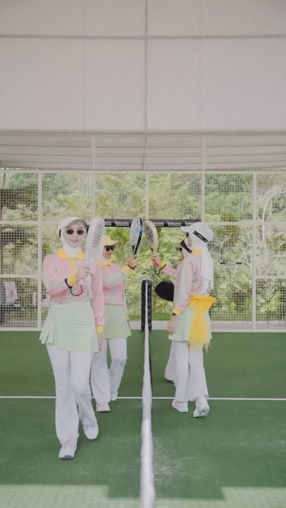 wanita bermain padel tos raket di lapangan tenis, mengenakan pakaian formal berwarna pastel.