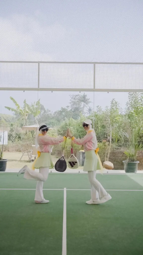 wanita bermain padel berpose tos di lapangan tenis, pakaian seragam, latar belakang hijau.