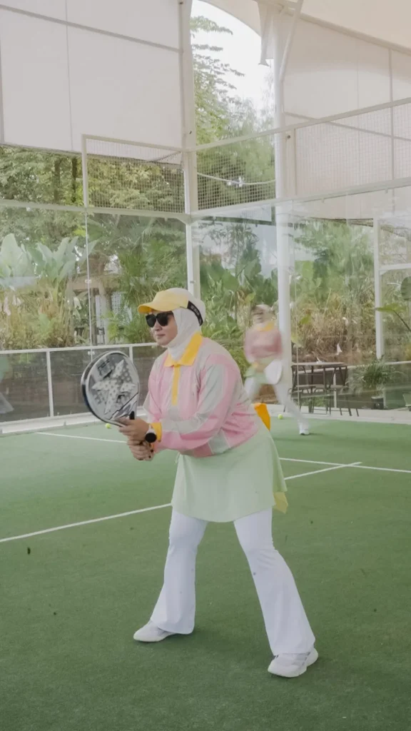 wanita bermain padel berpose di lapangan tenis, pakaian warna pastel, latar belakang taman hijau.