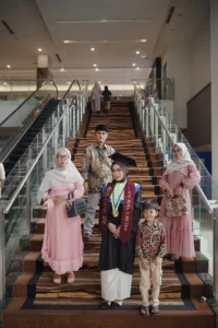 Seorang mahasiswa Aila Stikes Purwokerto Aston sedang berpose di atas tangga, menampilkan pakaian lulusan.