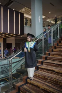 Aila dari Stikes Purwokerto Aston sedang berdiri di atas tangga, memakai sarung toga dan topi lulusan, mengambil foto untuk wisudanya.