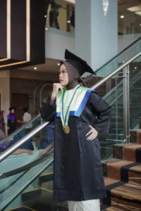 Aila Stikes Purwokerto Aston dalam foto wisuda mereka berdiri di depan escalator dalam pakaian sarjana.