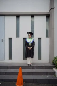 Aila Stikes Purwokerto Aston dalam foto wisuda berpakaian sarjana berdiri di depan gedung.