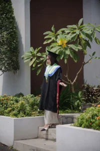 Aila Stikes Purwokerto Aston dalam foto wisuda berdiri di depan gedung.