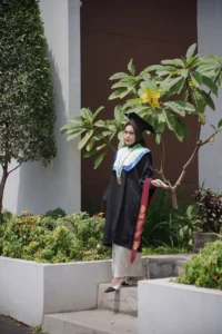Aila Stikes Purwokerto Aston, seorang wanita dalam pakaian wisuda, berdiri di depan pohon.