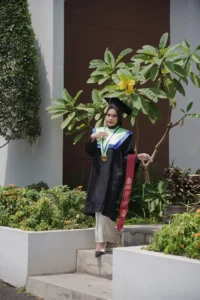 Aila Stikes Purwokerto Aston, seorang wanita dalam pakaian sarjana berdiri di depan pohon, membawa gelar sarjana.