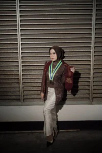 Aila, seorang mahasiswa Stikes Purwokerto Aston, sedang berpose di sekitar pagar, memakai dress elegan dan menampilkan medali prestasi.