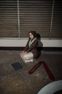 Ayla Stikes Purwokerto Aston sedang berwisuda di atas tanah, memakai pakaian gelap dan membawa objek biru.