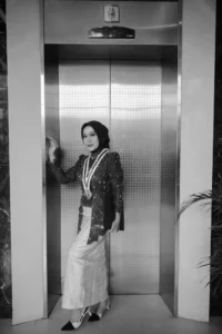 Aila Stikes Purwokerto Aston di foto wisudanya sedang berdiri dalam lift, memakai pakaian formal.