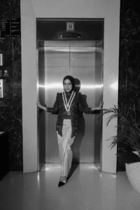 Aila Stikes Purwokerto Aston dalam foto wisudanya berdiri di lift, memakai pakaian hitam dan putih sambil berpose dengan tangan di pinggul.