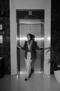 Aila Stikes Purwokerto Aston, seorang wanita berpakaian hitam putih, berdiri di dalam lift, menunjukkan pose dengan tangan di pinggang.