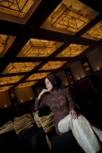 Aila Stikes Purwokerto Aston sedang berfoto wisuda di ruangan dengan pola langit, sementara dia berdiri dengan tangan di pinggul.