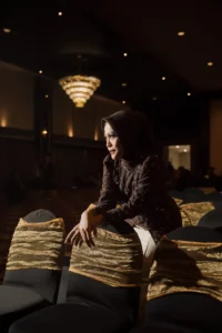 Seorang wanita dalam pakaian hitam duduk di kursi dengan tangan di bawah dada, memandang samping, foto wisuda Aila Stikes Purwokerto Aston.