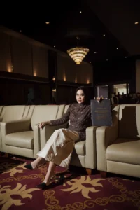Aila Stikes Purwokerto Aston sedang berwisuda di tengah foto yang menampilkan wanita duduk di sofa, membaca buku, dan tersenyum.