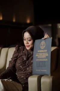 Aila Stikes Purwokerto Aston duduk di kursi, menahan gelar wisuda yang diterimanya.