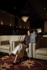 Aila Stikes Purwokerto Aston sedang menikmati momen wisuda di ruang tamu, duduk di sofa sambil membaca buku.