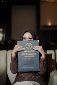 Aila Stikes Purwokerto Aston sedang mengambil foto wisuda sambil membawa buku, tersenyum, dan berpose untuk kamera.