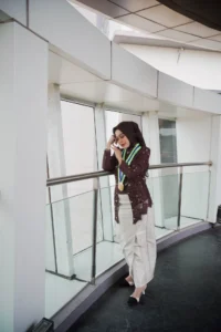 Aila Stikes Purwokerto Aston sedang berwisuda di atas balconya, memegang telepon di telinganya sambil memakai pakaian putih.