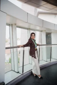 Aila Stikes Purwokerto Aston dalam foto wisudanya berdiri di atas tangga dengan pakaian putih dan hitam, tangan di atas balustrade.