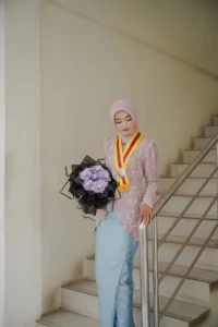 Kristina, seorang mahasiswa Universitas Muhammadiyah Purwokerto, sedang memperingati wisudanya dengan memakai pakaian berwarna biru dan membawa bunga-bunga, berdiri di atas tangga.