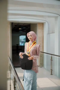 Kristina UMP, seorang wanita berpakaian formal, berdiri di depan gedung Universitas Muhammadiyah Purwokerto sementara membawa tas.