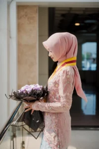 Kristina UMP universitas Muhammadiyah Purwokerto dalam foto wisudanya berdiri di depan gedung dengan bunga di tangan, memakai pakaian berwarna pink.