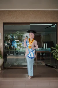 Seorang wanita dalam pakaian berwarna merah dan biru, memakai topi sarjana dan membawa gelar sarjana, sedang melalui pintu berbentuk kaca di Universitas Muhammadiyah Purwokerto.