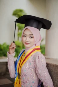 Kristina, seorang mahasiswa Universitas Muhammadiyah Purwokerto, sedang berpose untuk foto wisuda, memakai cap dan gowns lulusan serta membawa paras.