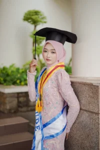 Kristina UMP, seorang mahasiswa Universitas Muhammadiyah Purwokerto, sedang berpose dalam pakaian lulusan sambil membawa sebuah伞.