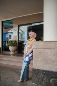 Seorang wanita dalam pakaian wisuda berdiri di depan dinding.