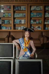 Kristina, seorang mahasiswa Universitas Muhammadiyah Purwokerto, sedang duduk di bangku dengan gelang sastra di lehernya, menyelesaikan studinya dengan pakaian sarjana.