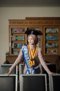 Kristina UMP, seorang wanita dalam cap dan gowns wisuda, berdiri di depan rak buku.