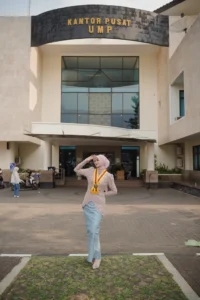 Kristina UMP, seorang mahasiswa Universitas Muhammadiyah Purwokerto, berdiri di depan gedung, memakai pakaian berwarna pink dan merah, dan melihat sisi kiri.