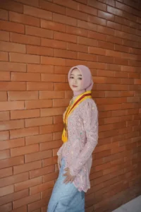 Kristina UMP, seorang mahasiswa Universitas Muhammadiyah Purwokerto, berdiri di depan dinding bata, memakai baju berwarna pink dan putih serta kerudung berwarna merah.