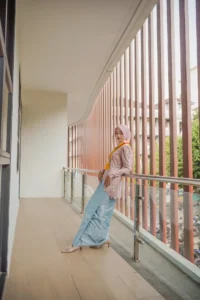Kristina, seorang mahasiswa Universitas Muhammadiyah Purwokerto, sedang berdiri di atas balcony, memakai top berwarna pink dan jeans biru, dengan wajah terbuka sementara melihat sisi.