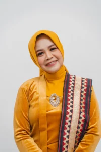 Seorang wanita berhijab berdiri di depan kamera, memakai pakaian berwarna kuning dan sedang mengambil foto.