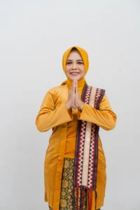 Seorang wanita berdiri dengan tangan bersama, memakai pakaian kuning dan headscarf, di foto profil kepala Kementerian Kebudayaan dan Pariwisata Provinsi Purbalingga.
