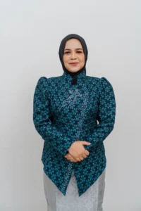 Seorang wanita berdiri dengan tangan bersama, memakai pakaian berbordir biru dan hitam, di depan Kepala Kementerian Kebudayaan dan Pariwisata Provinsi Purbalingga.