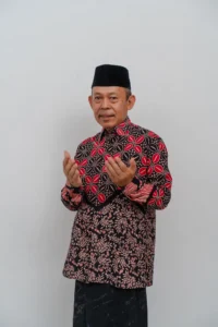 Seorang pemimpin kementerian dalam pakaian tradisional, dengan tangan terbuka di depan, berdiri di latar belakang putih.