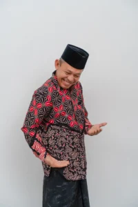 Seorang lansia berpakaian tradisional Indonesia berdiri dengan tangan di pinggangnya.