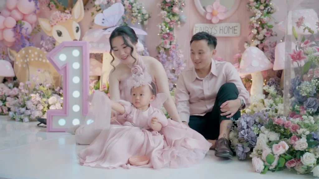 Keluarga berpose dengan bayi di depan latar belakang tema pernikahan, semua berpakaian pink.