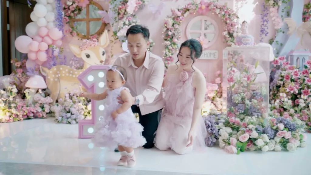 Anak perempuan berjalan di depan ayah dan ibu, berpakaian cantik, di depan dekorasi berwarna pink dan